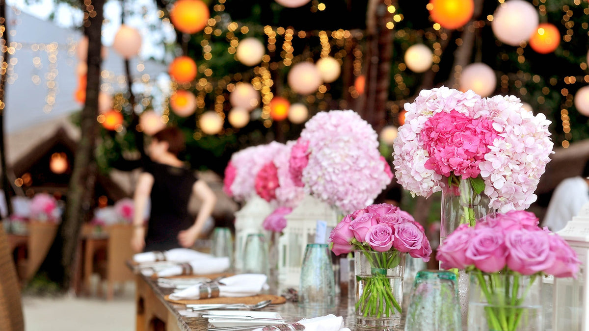 An outdoor evening reception table set with pink hydrangeas and roses, beneath paper lanterns and string lights.